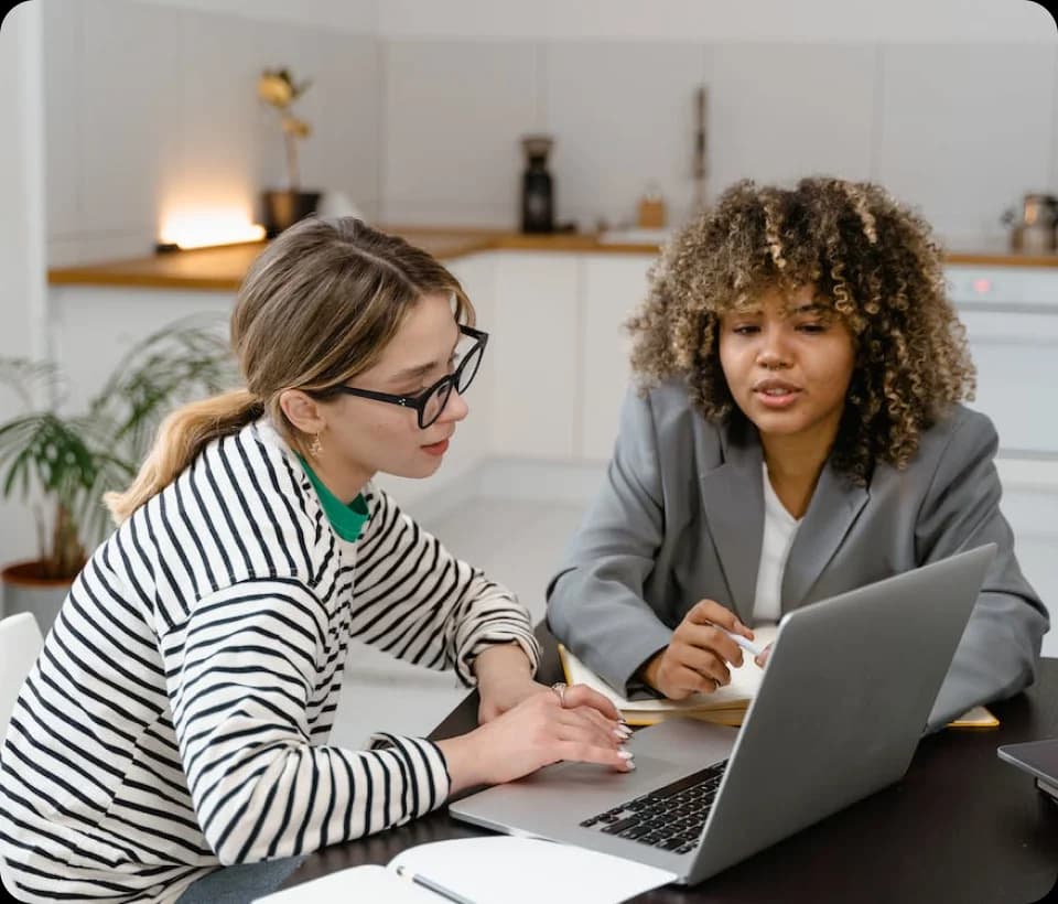 Zwei Frauen sitzen an einem Tisch und schauen gemeinsam auf einen Laptop, während sie in einem modernen Raum diskutieren.